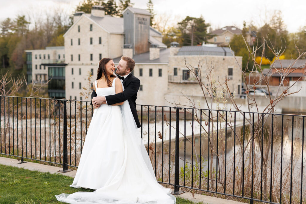 Bride and groom embracing at Elora Mill with riverside views.