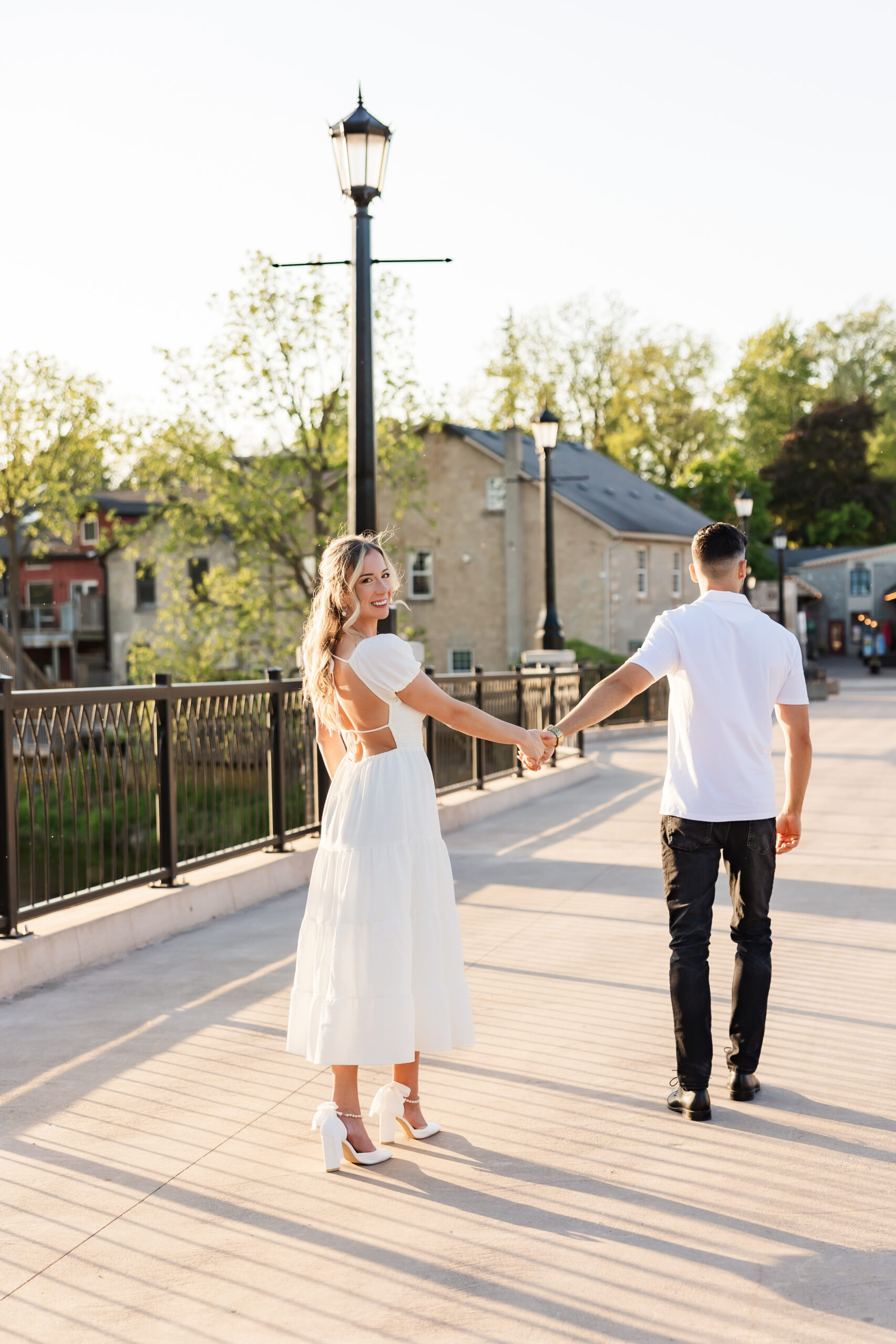 Bride-to-be in white dress holding hands with fiancé across Elora bridge at sunset, smiling back at the camera